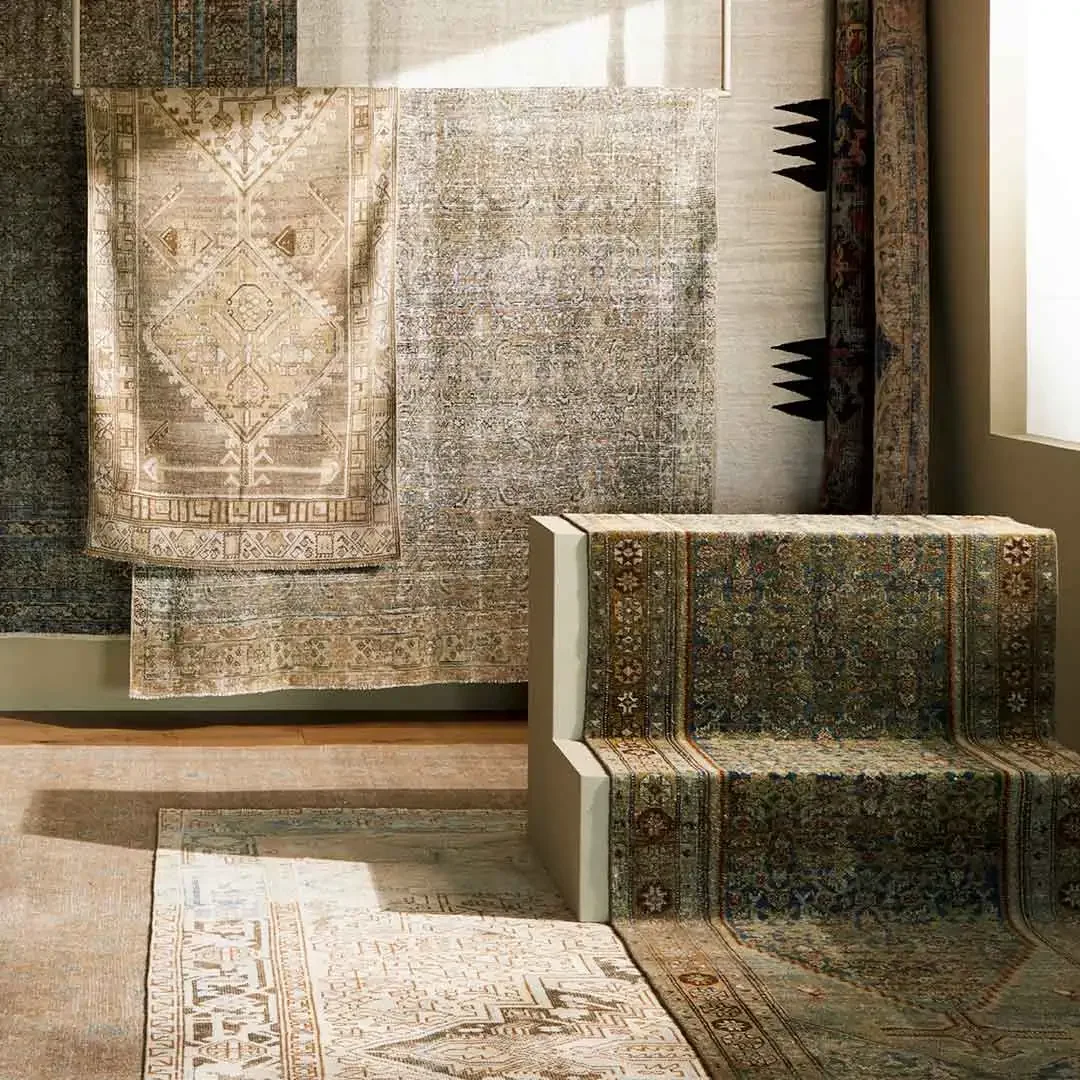Multiple Surya designer unique new and vintage patterned rugs hanging on the wall and laid on the floor, with a small bench or seat covered in a similar one-of-a-kind rug, inside a room with a window.