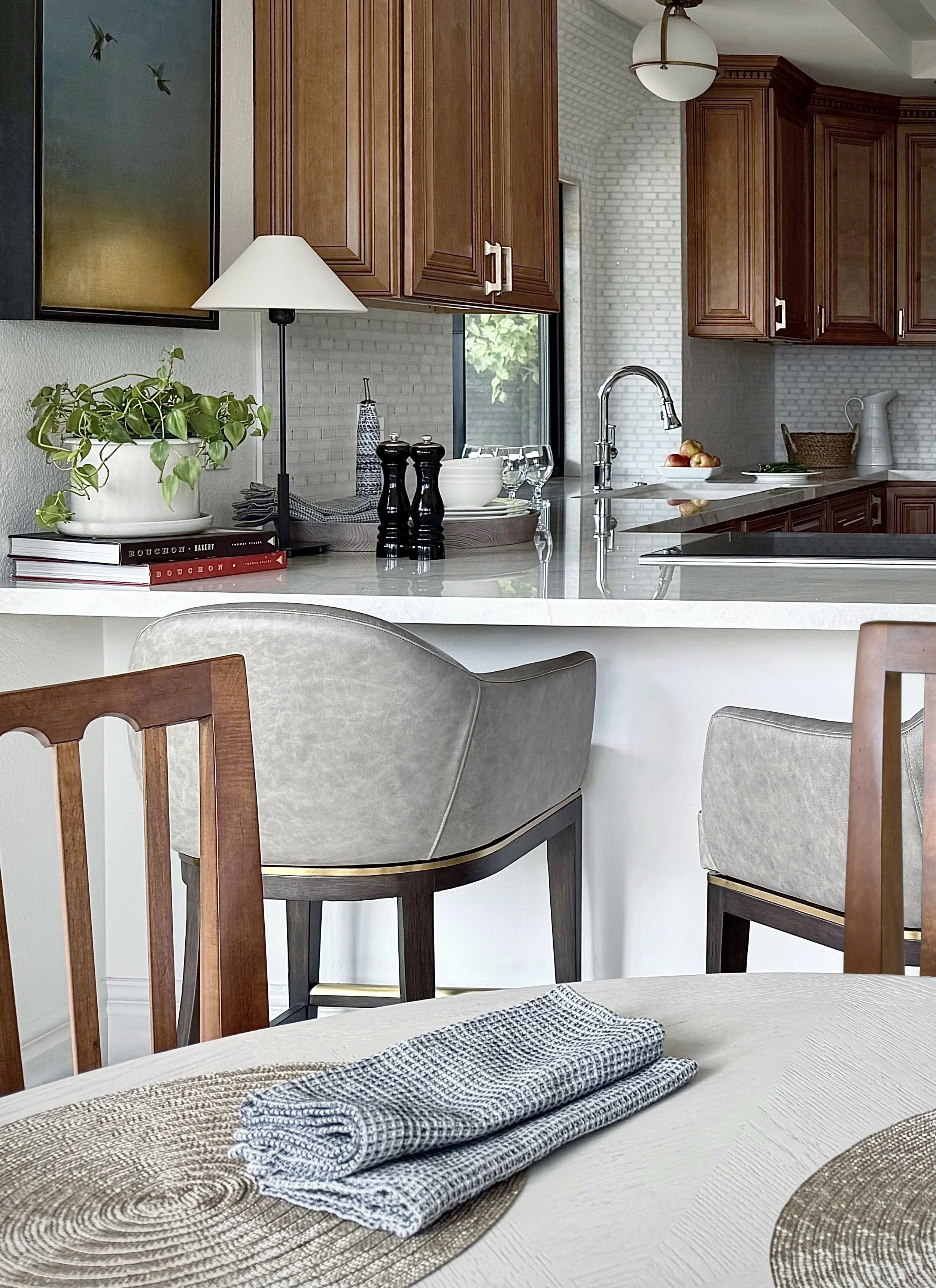 Rancho Mirage designer kitchen with birch cabinets, Taj Mahal quartzite countertop, leather counter stools. Items on the counter include pepper mill, bowls, lamp. There is a globe light fixture and a marble-tiled backsplash window in the background.