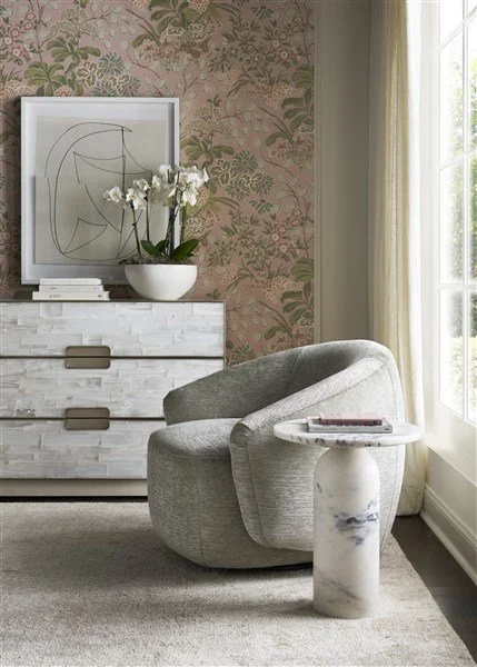A cozy corner with gray Vanguard armchair, round marble side table, white dresser with a framed abstract art piece, white orchid plant, and window with cream-colored curtains letting in natural light, all against a pink and green botanical wallpaper.