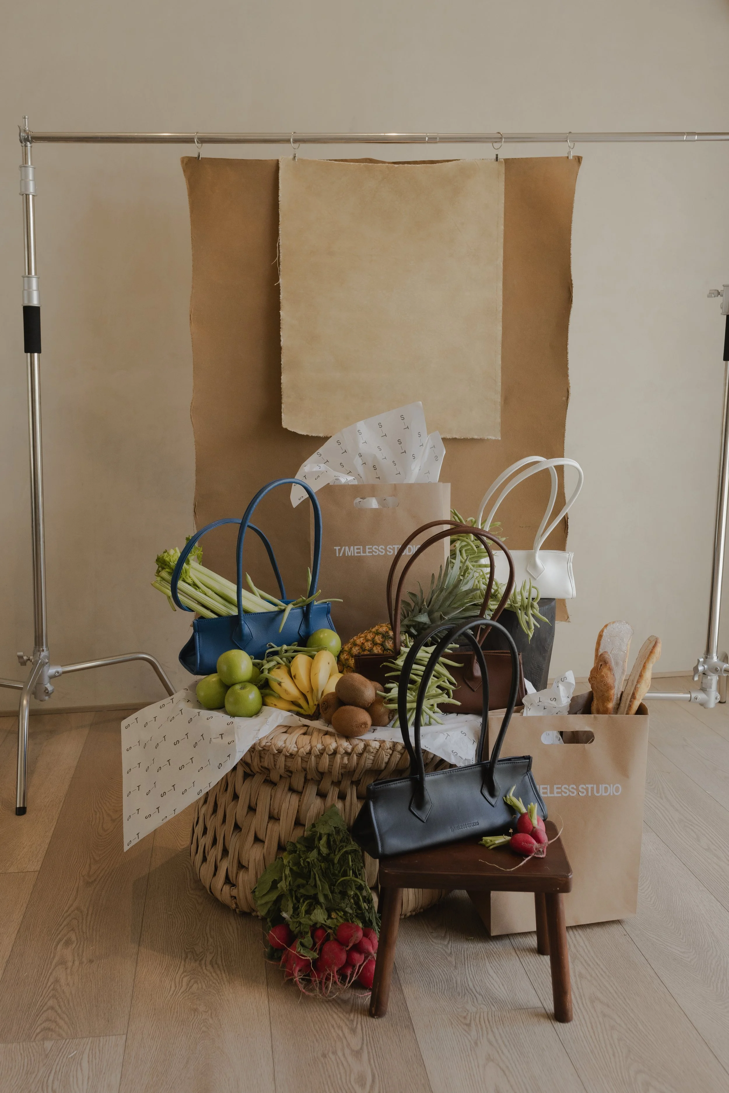 Variedad de frutas, verduras y bolsas de compras en una presentación de estilo de mercado en interior, con fondo de tela y soporte metálico.
