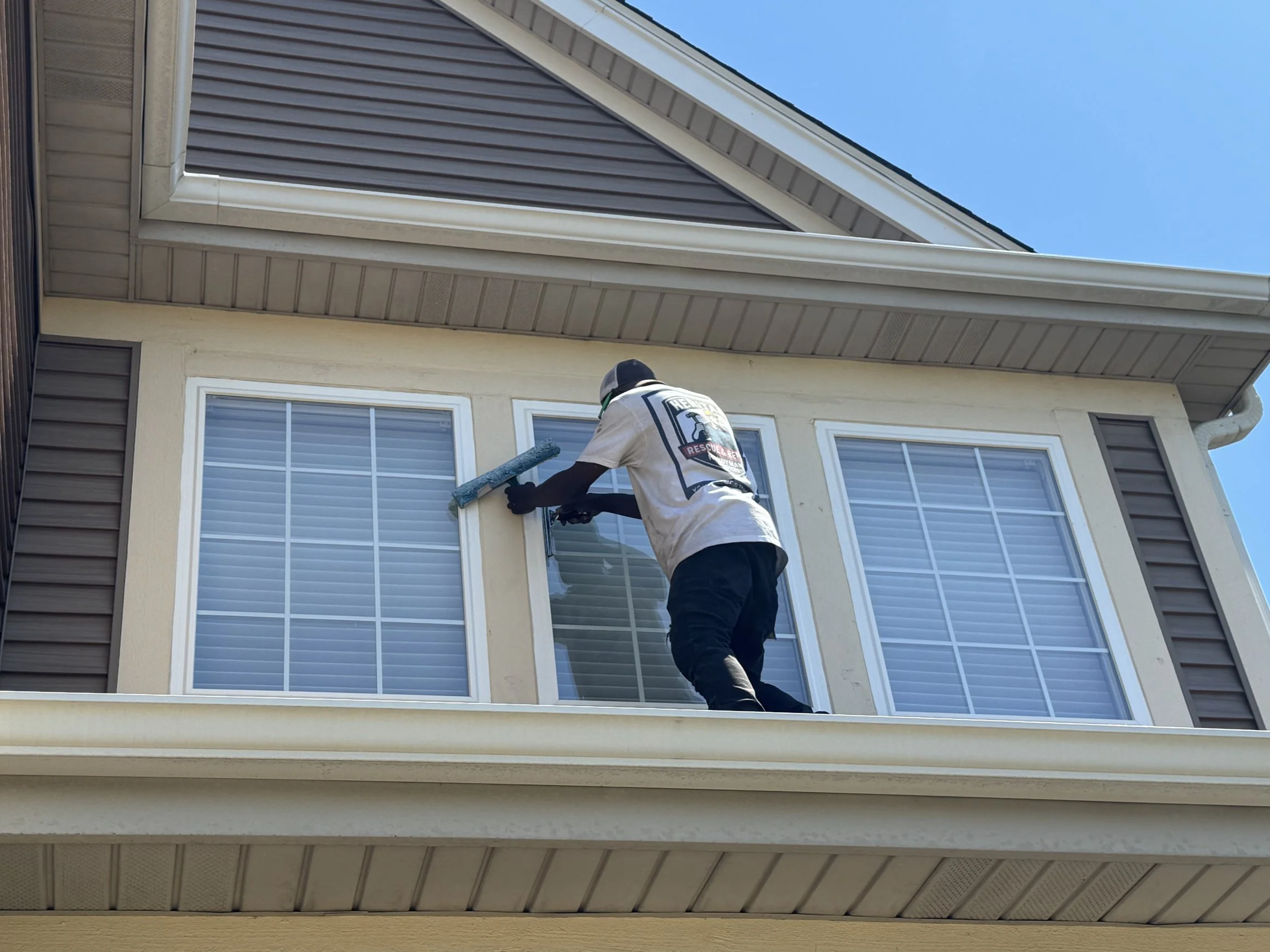 A worker on a ladder cleaning the exterior window of a two-story house with blond siding, white trim, and double-pane windows, against a clear blue sky.