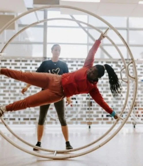 A woman in a red jacket performing an acrobatic pose inside a large circular metal ring, with a man standing behind her in a spacious, well-lit gym or studio.