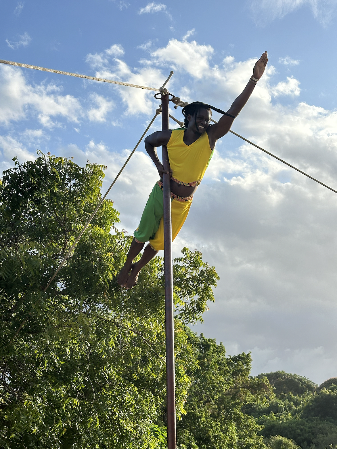 A man balancing on a tightrope made of a pole and ropes, smiling, during daytime with clouds and trees in the background.