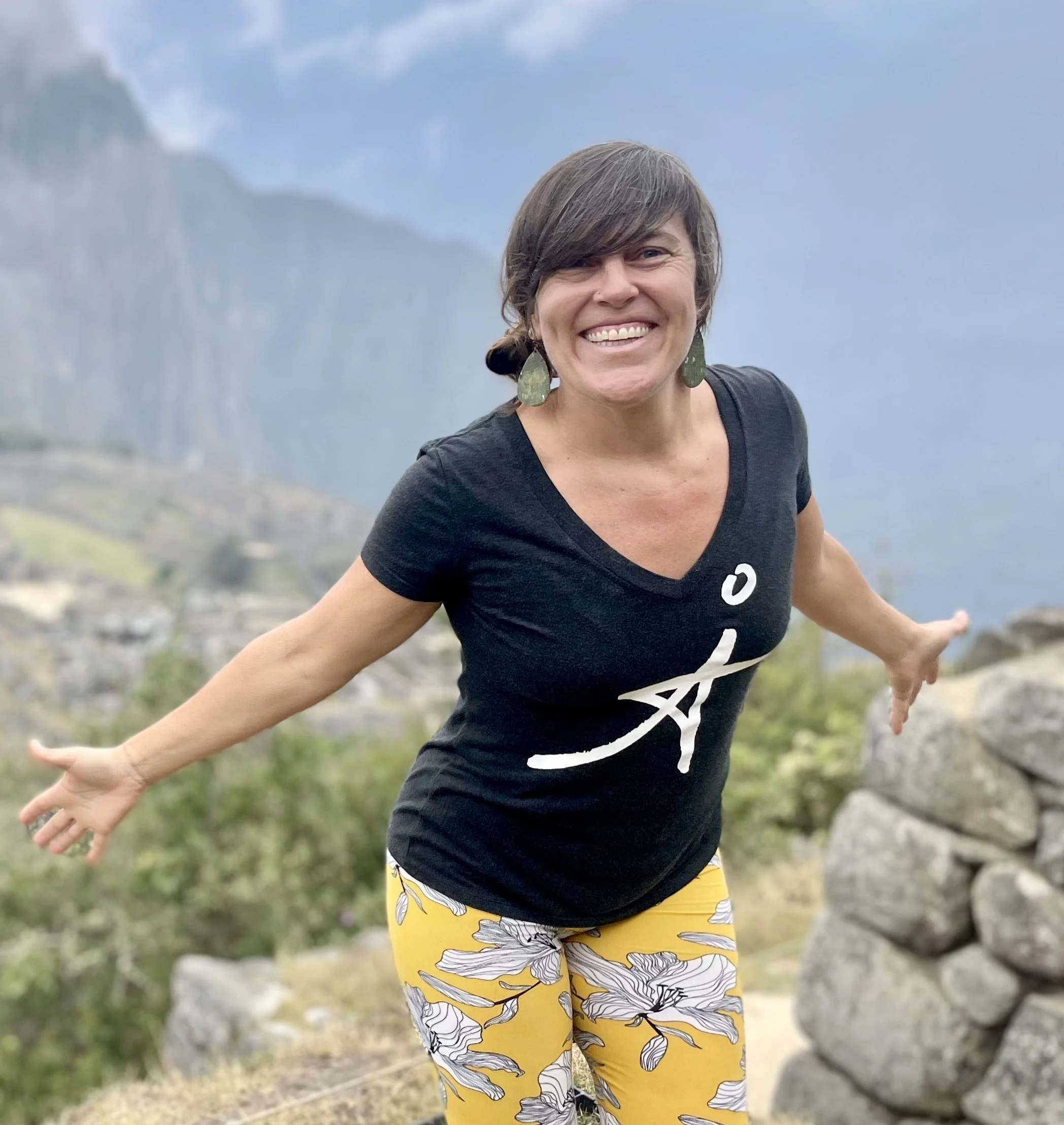 A woman smiling with arms outstretched in a mountainous outdoor setting, wearing a black T-shirt and yellow floral pants.