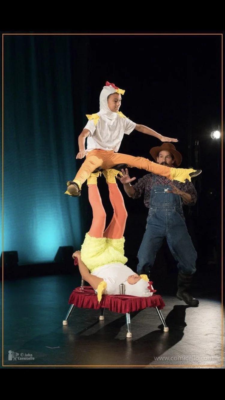 A person in a chicken costume balancing on a table, with two performers supporting them from below, while a third performer stands nearby.