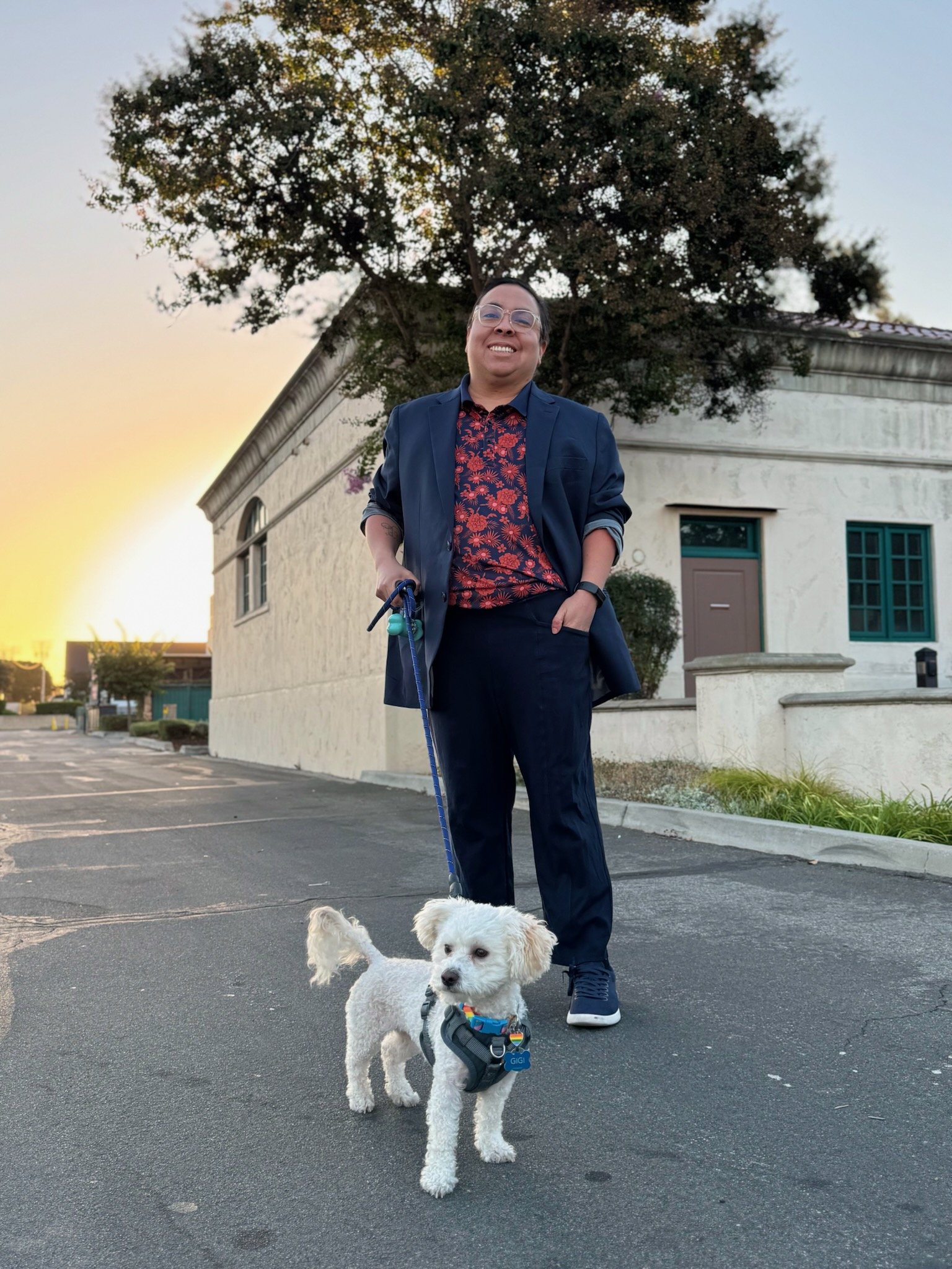 A person standing with a flower button up collared shirt holding a leash and smiling. He is holding a leash that connects to his white poodle mix dog Gigi who is looking out in the distance.