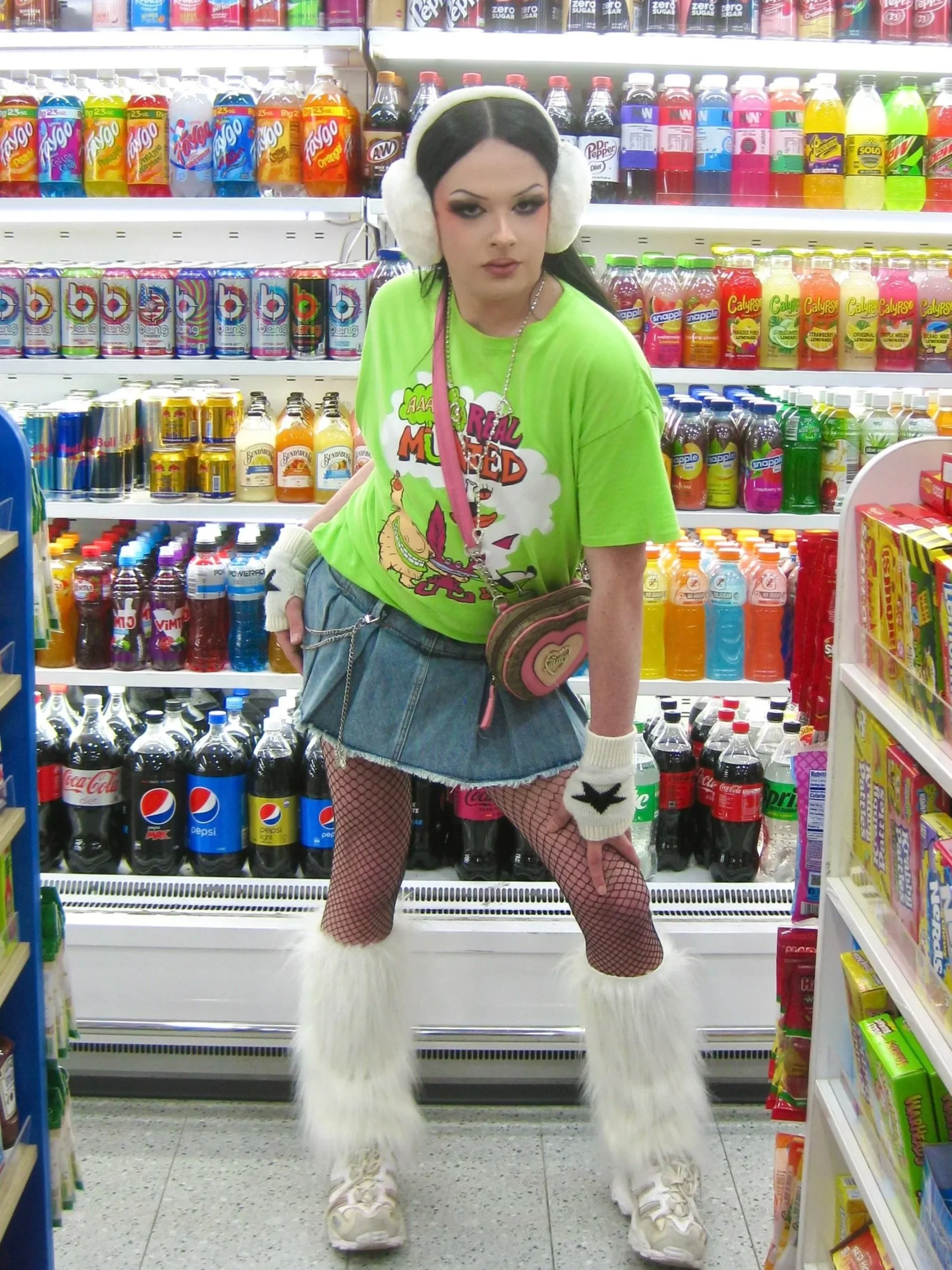 A young woman with dark hair and makeup in a colorful, eclectic outfit shopping at a convenience store aisle filled with soda bottles.