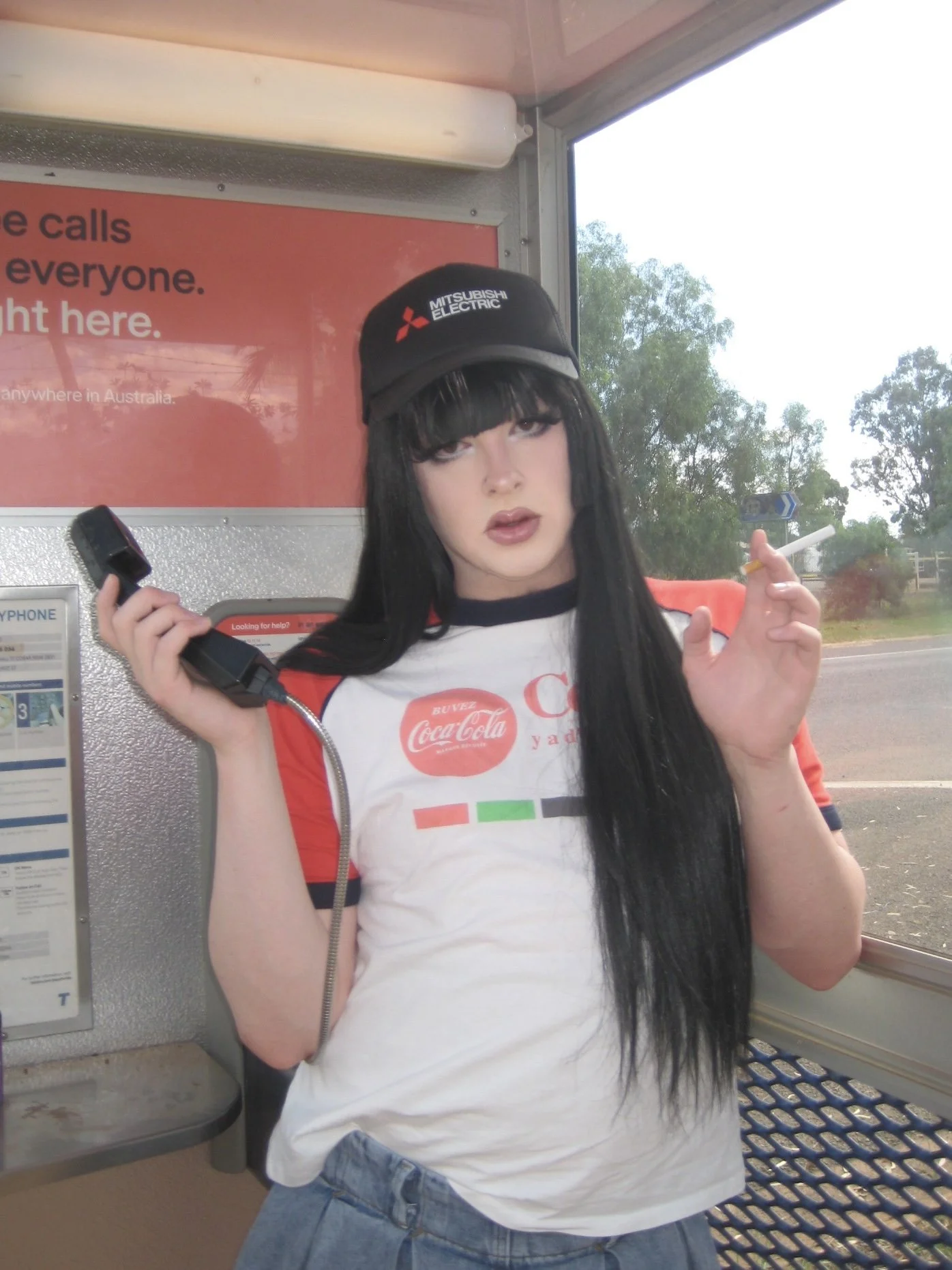 A young woman with long black hair and bangs standing at a bus stop, holding a cigarette in one hand and a phone in the other. She is wearing a black cap with the Mitsubishi Electric logo and a white and red Coca-Cola t-shirt.