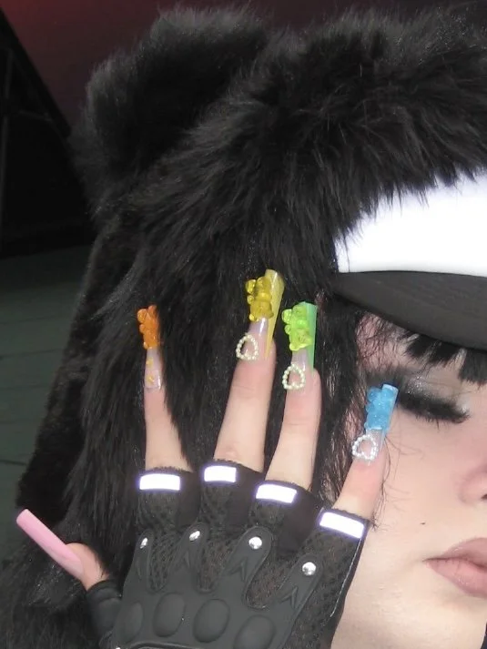 A person with colorful, cactus-shaped rings on each finger, wearing black gloves with reflective strips, holding manicured nails with decorative rings, and wearing a black furry hat and a visor.