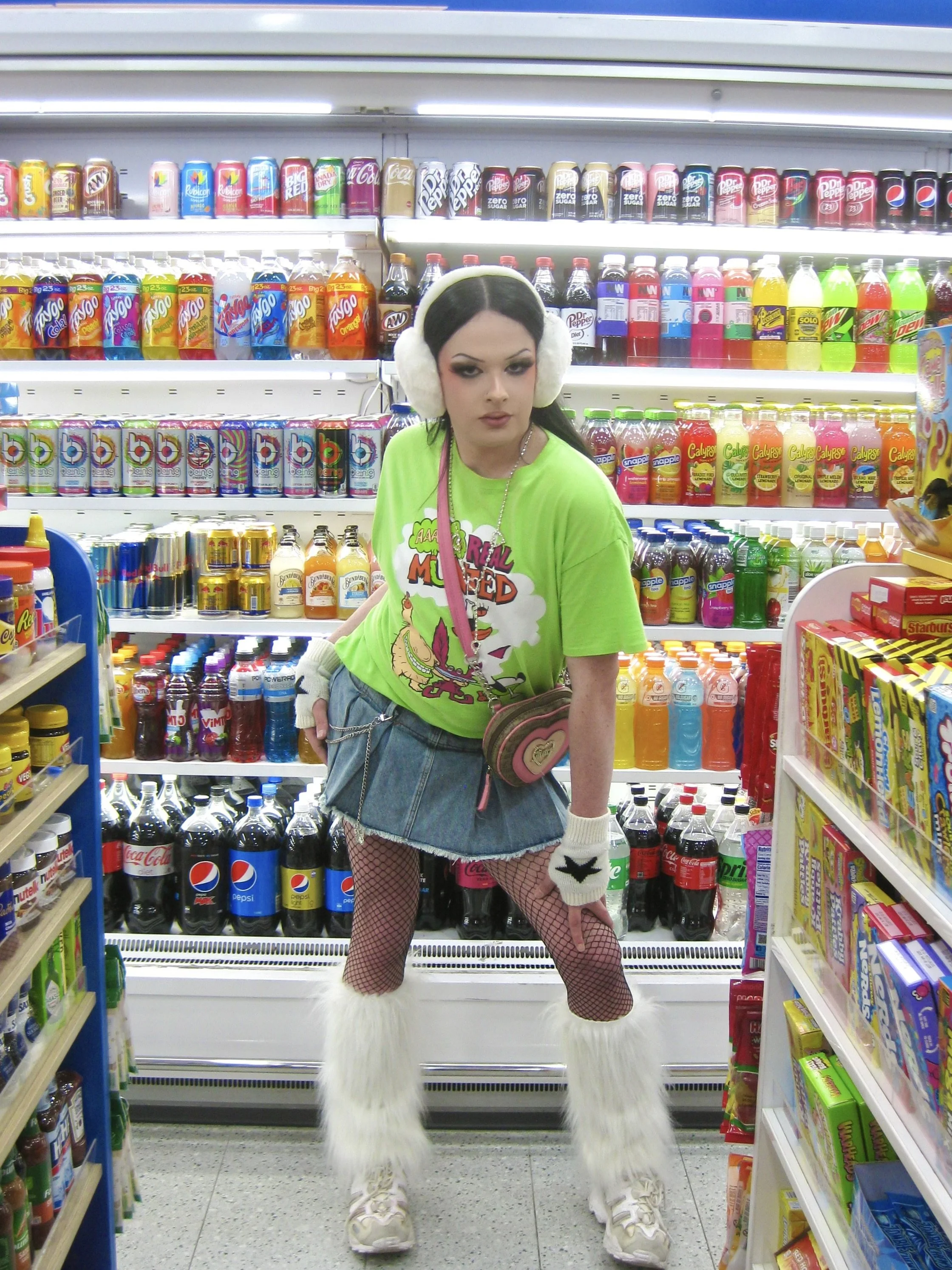 A young woman with black hair, wearing white fluffy earmuffs, a bright green T-shirt with illustrations, a denim skirt, fishnet stockings, furry white boots, and white gloves with black star designs, standing in a grocery store aisle with colorful drinks on shelves behind her.