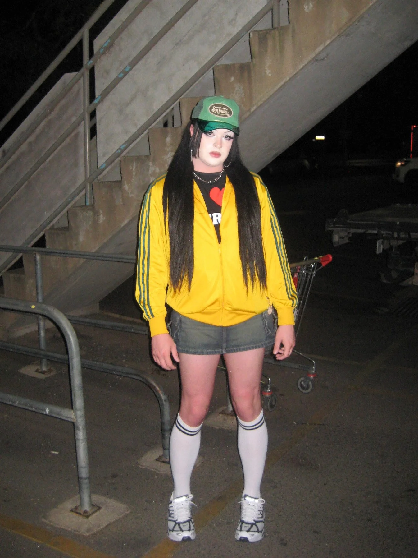 A woman with long black hair wearing a green cap, yellow jacket, short gray skirt, white knee-high socks, and sneakers standing outdoors at night near a staircase, with a shopping cart in the background.