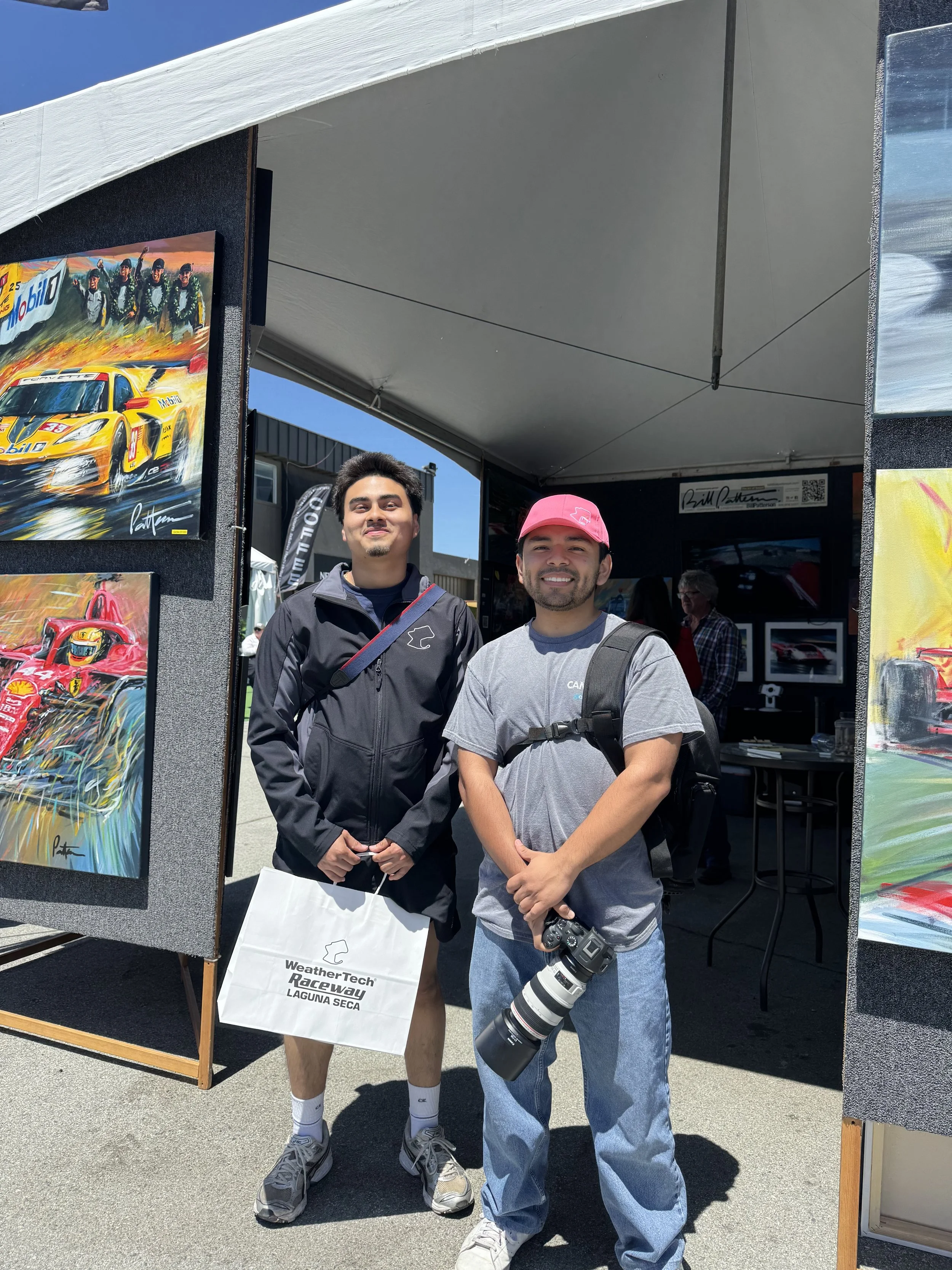 Two men standing in front of race car artwork at an outdoor event, one holding a camera with a large telephoto lens.
