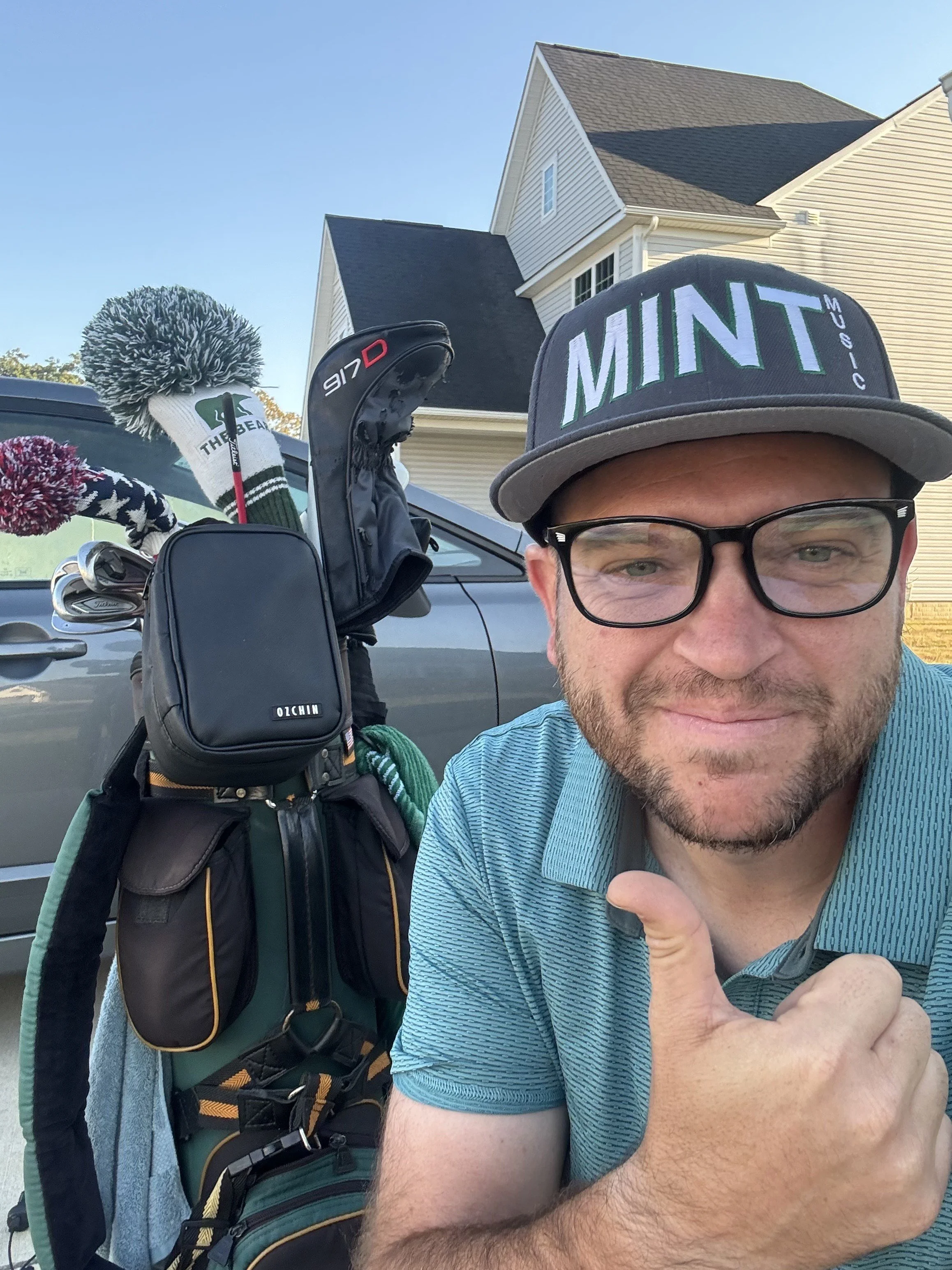 A man with glasses and a cap that says MINT is taking a selfie outdoors near a golf bag with clubs and headcovers. There are houses and a clear sky in the background.