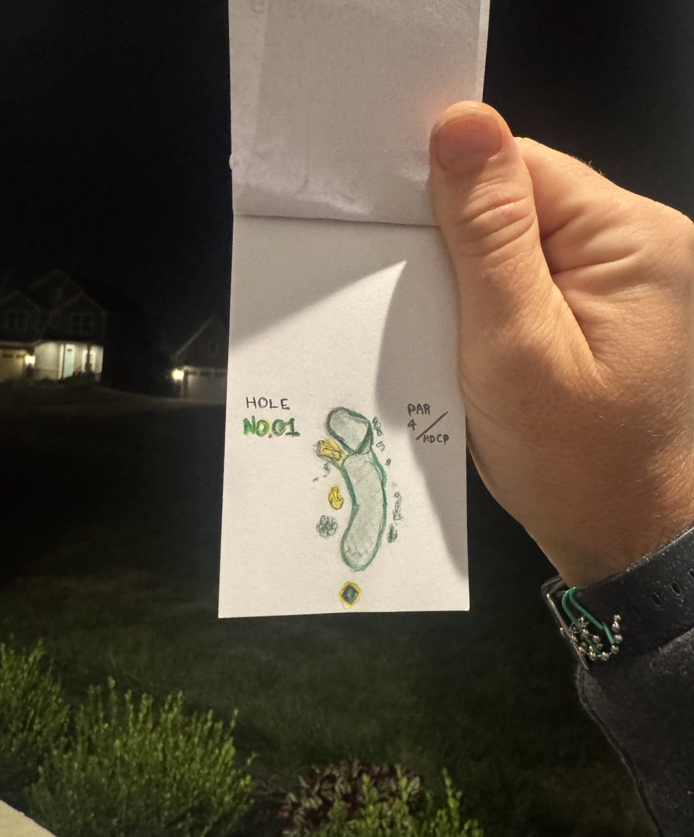 Hand holding a small paper golf course map at night, showing a hole marked NO. 1 with landscaping and a par 4, adjacent to a dark residential neighborhood.