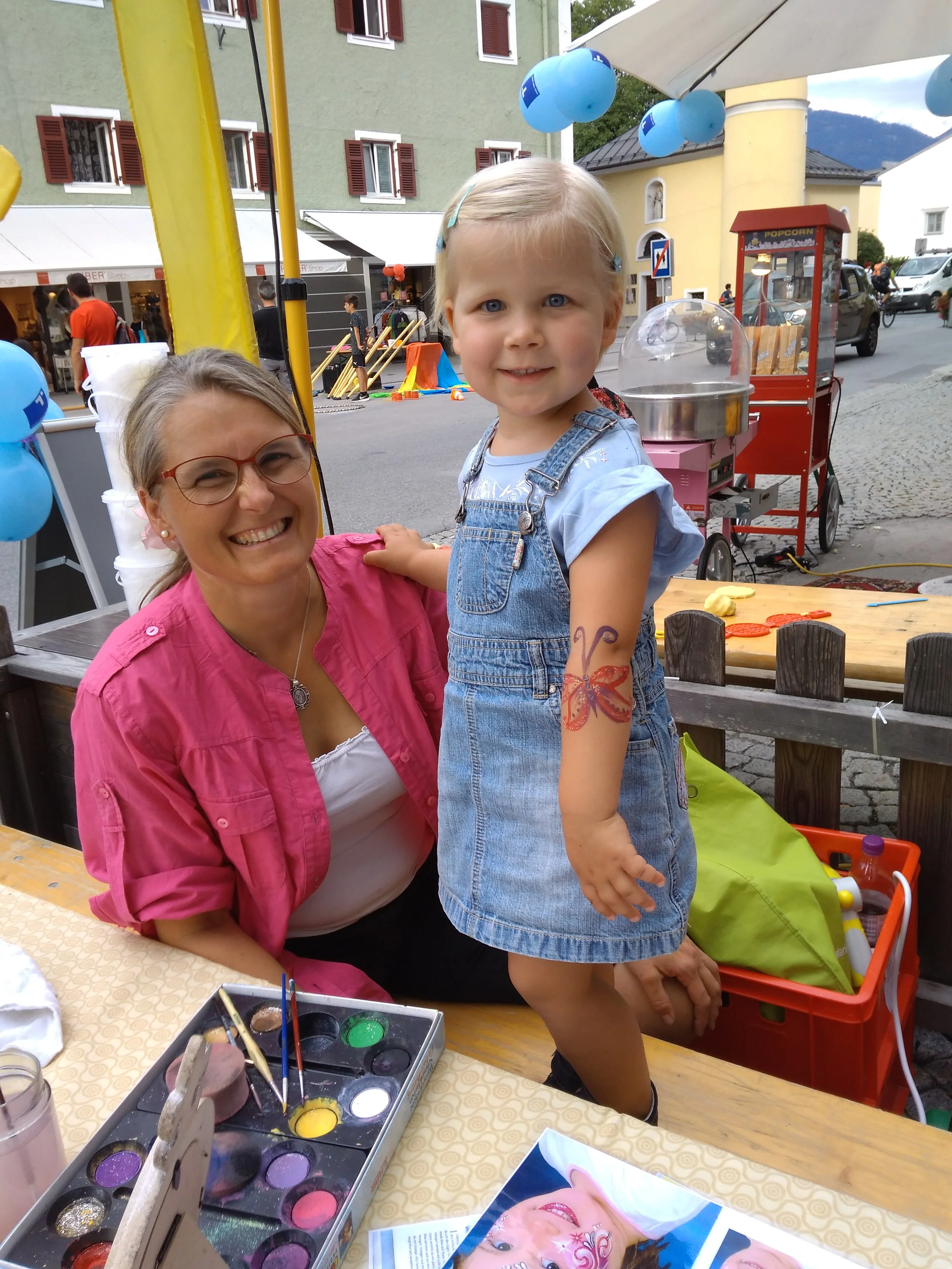 Eine Frau und ein kleines Mädchen beim Schminken mit bunten Farben auf einem Tisch, draußen auf einer Straße mit bunten Gebäuden und Volksfestdekoration.
