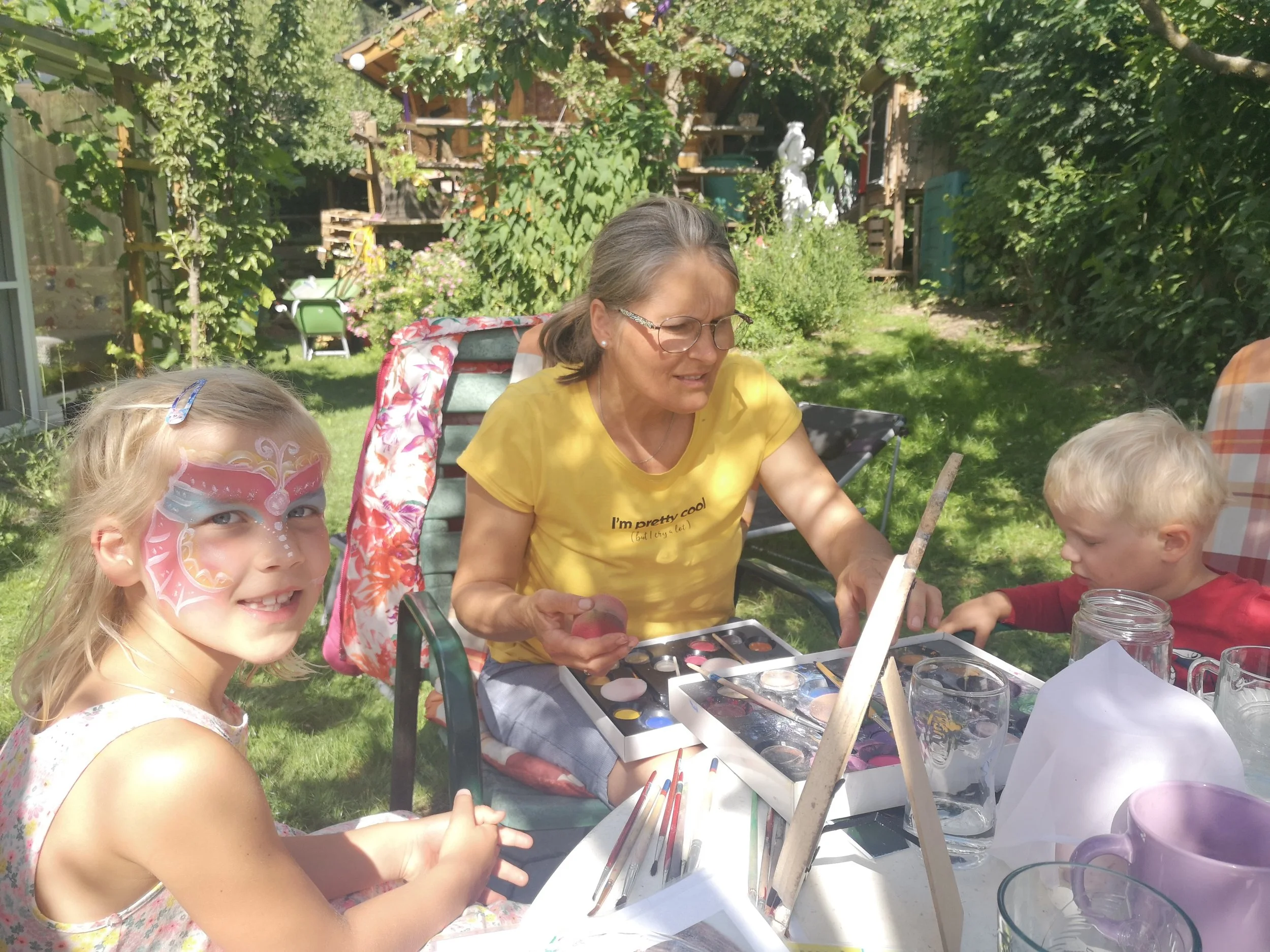 Eine Frau macht Gesichtsbemalung und Kinderschminken für ein Mädchen mit einem Schmetterlingsmotiv im Garten, während ein Junge zuschaut und auf dem Tisch Malutensilien liegen.