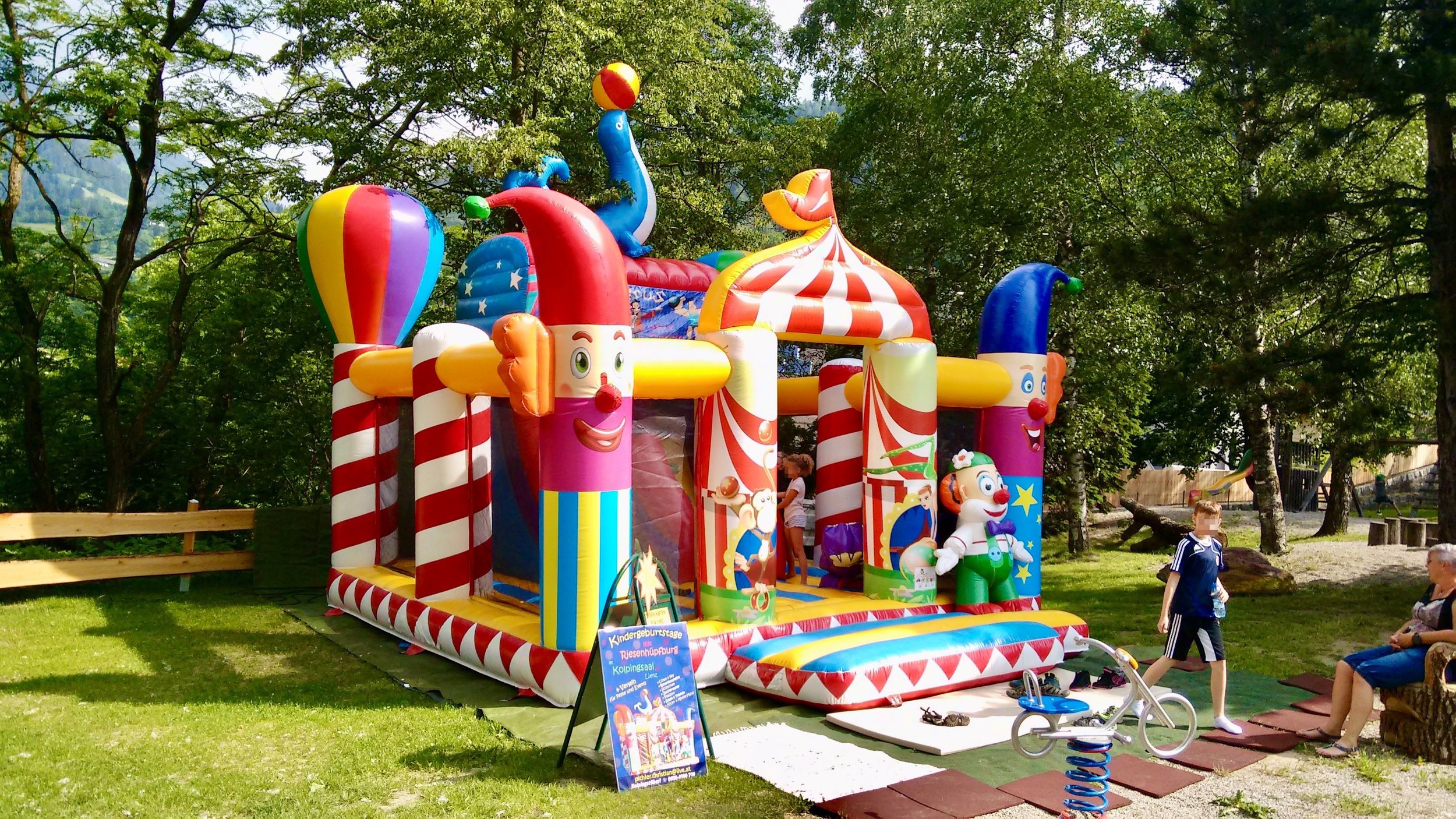 Bunte Zirkus-Hüpfburg mit Zirkusmotiven in einem Park, umgeben von Bäumen und Familien