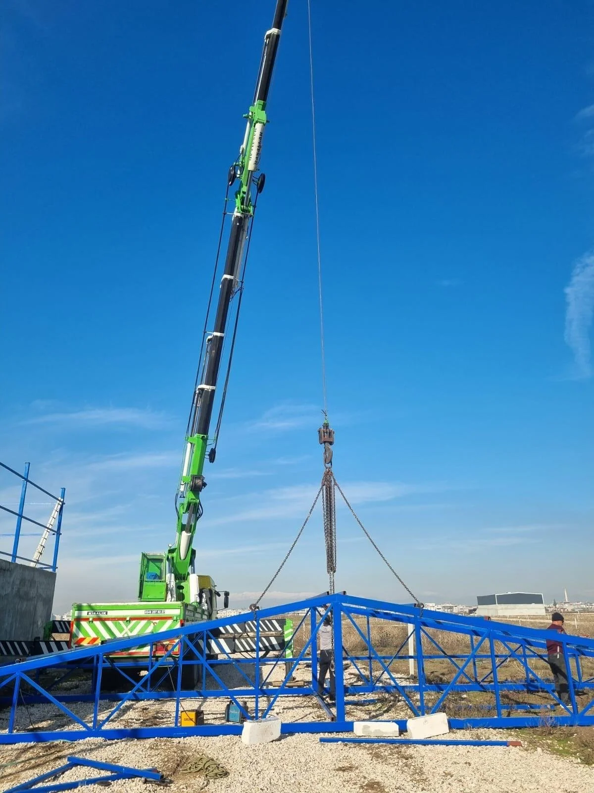Construction crane lifting a blue metal structure at a construction site under a clear blue sky.