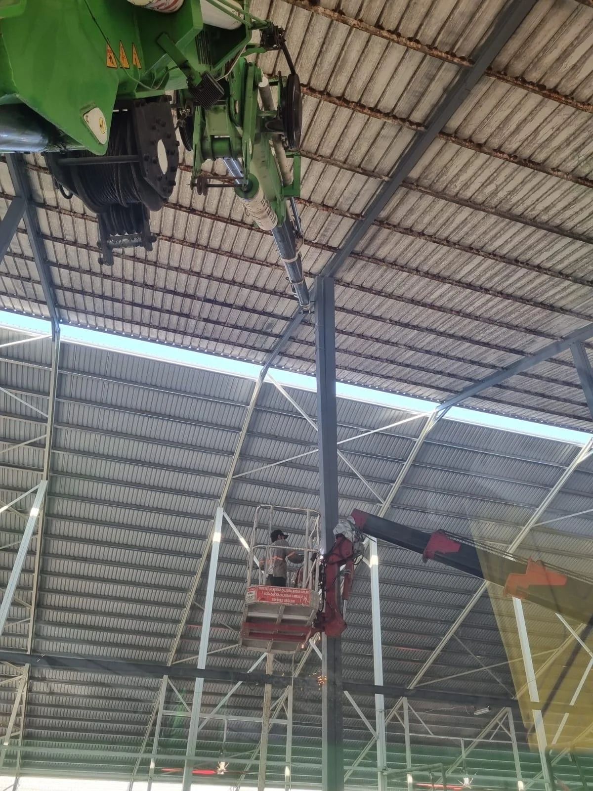 A worker on a cherry picker inspecting a large, green construction or industrial machine inside a warehouse with a metal roof.
