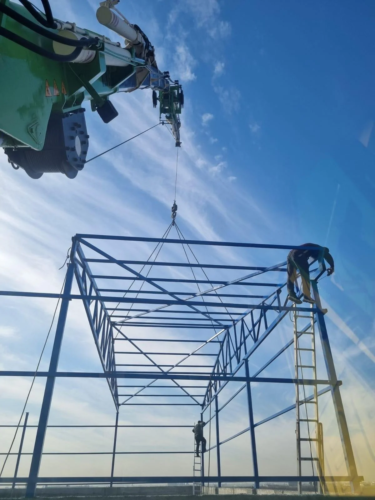 Workers assemble a large metal framework, with some climbing a ladder and others working at different heights, while a crane lifts and positions parts of the structure against a blue sky with scattered clouds.