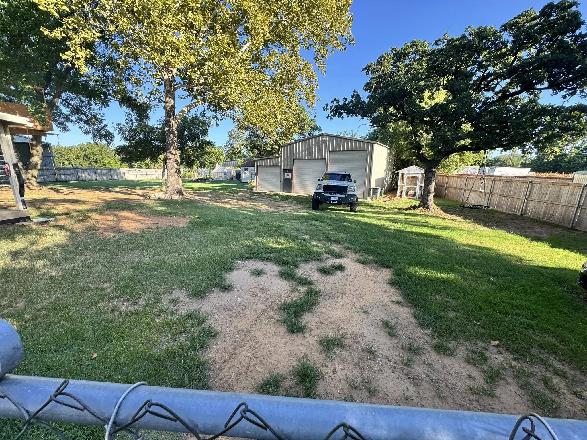 View of a backyard with grass, trees, a shed, a pickup truck, and a swing set, bordered by a wooden fence under a partly cloudy blue sky.