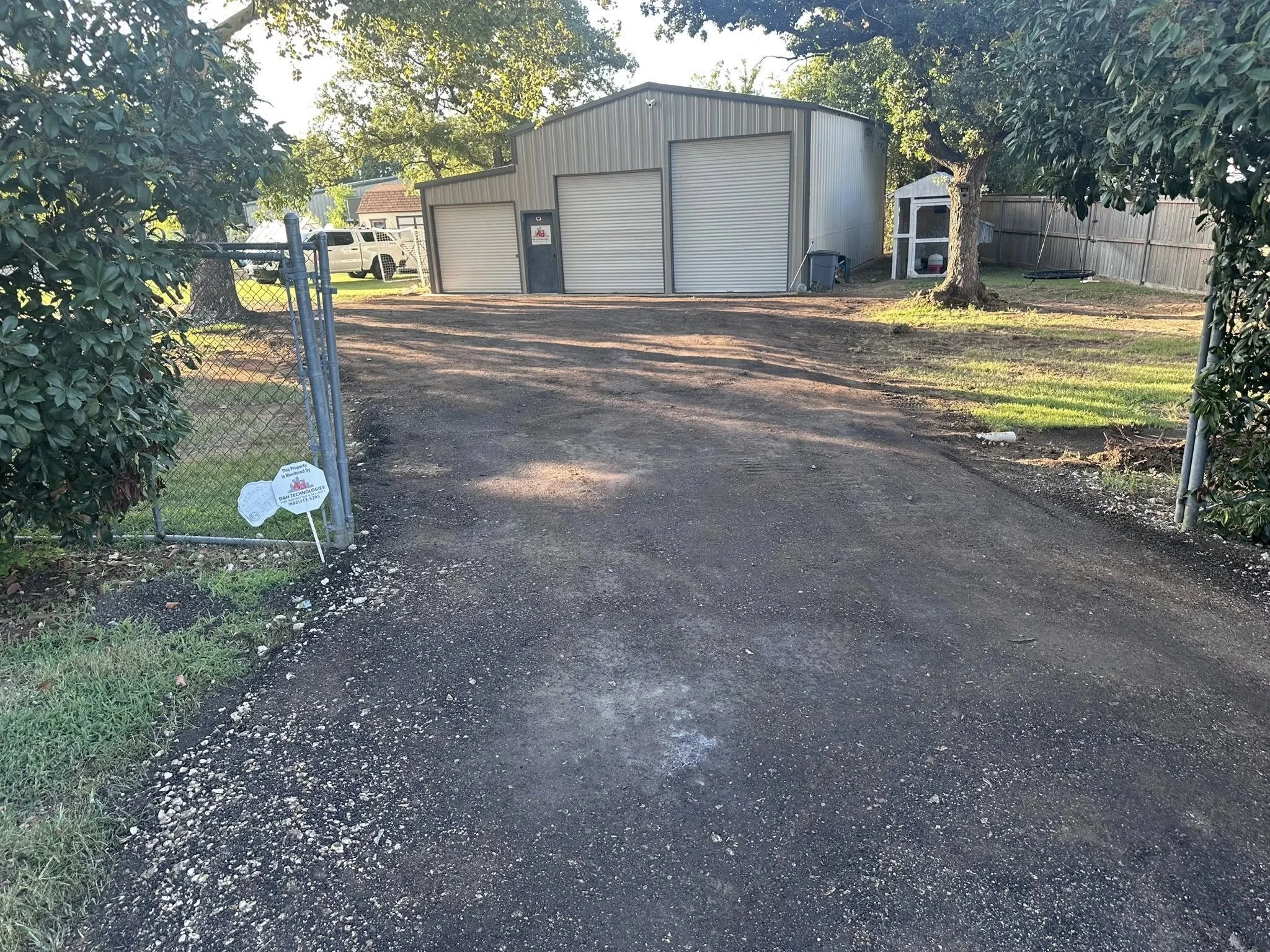 Gravel driveway leading to a green metal garage with three closed roll-up doors, trees on the sides, and a small shed in the backyard. A white vehicle is parked behind the gate, with a chain-link fence on the left. Sunlight creates shadows on the gro
