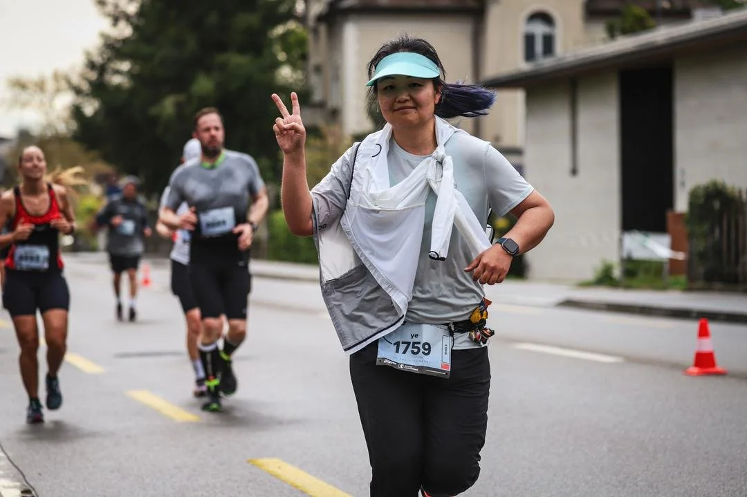 A woman running in a marathon, smiling and making a peace sign with her right hand, wearing a light blue visor and sportswear, with other runners behind her on a street.