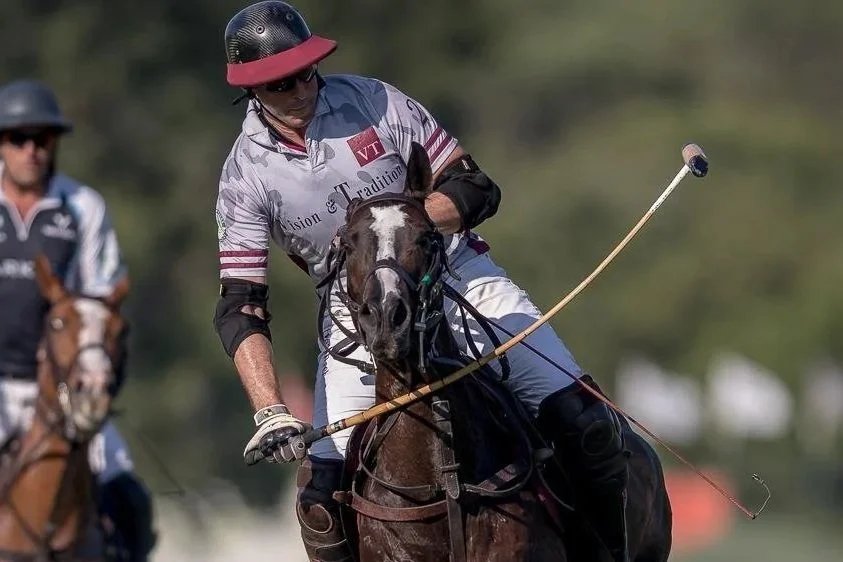 A polo player wearing a black helmet, sunglasses, and a white jersey riding a dark horse during a match, holding a mallet.