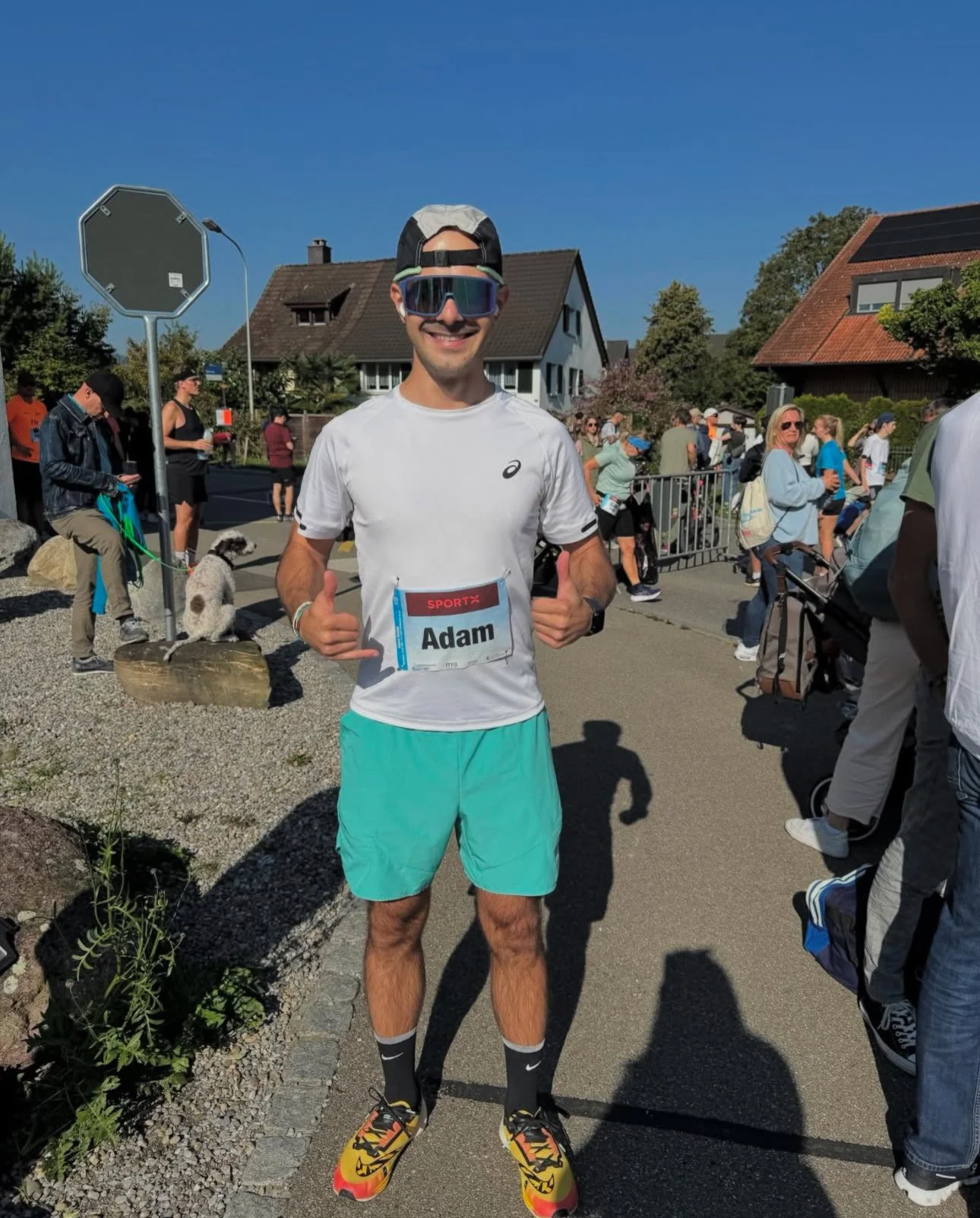 A male runner wearing sunglasses, a baseball cap backwards, and a white running shirt with a race bib that says 'Adam.' He is smiling and giving a thumbs-up at a race event with people and houses in the background.