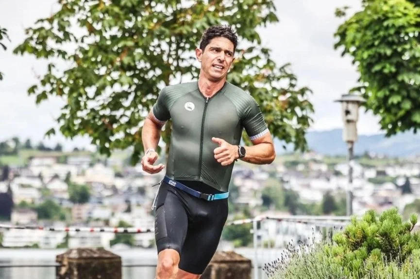 A man running outdoors in a park, wearing athletic gear and a smartwatch, with trees and a cityscape in the background.
