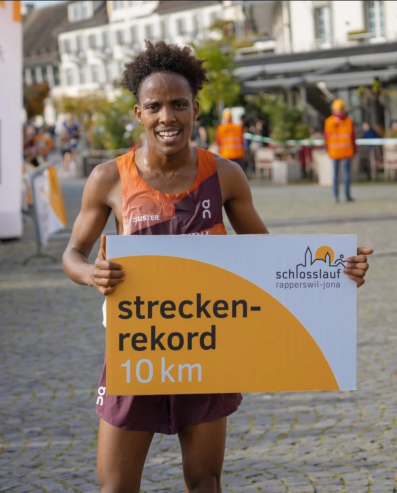 A smiling male runner holding a sign that reads "streckenrekord 10 km" at a finish line during a race event in an urban area.