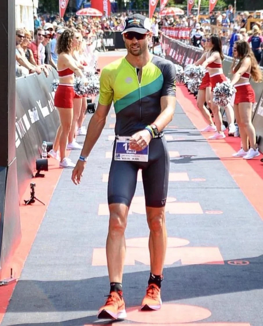 A male runner crossing the finish line at a race event, wearing a colorful athletic outfit, sunglasses, and a race bib, with cheerleaders dressed in red and white cheering on the sides on a sunny day.