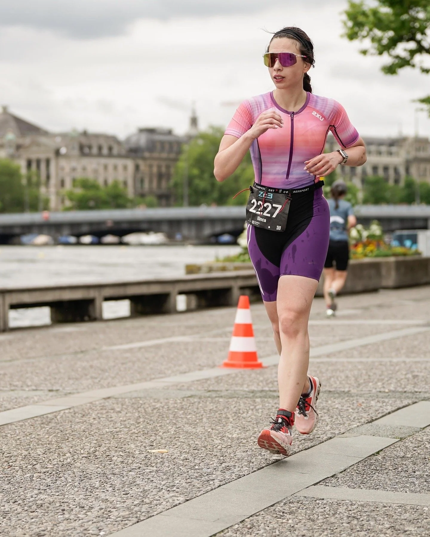 A woman running in a marathon, wearing sunglasses, a pink and purple athletic outfit, and a bib number 2227, on a paved path by a river with historic buildings and a bridge in the background, and an orange traffic cone nearby.