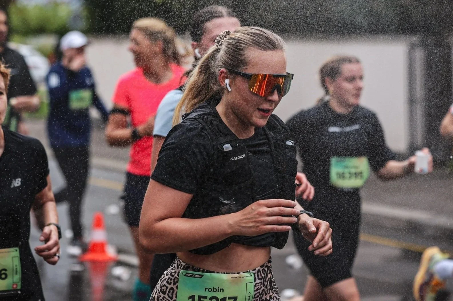 Female marathon runner, wearing sunglasses and wireless earbuds, runs in the rain among other runners, some with race bibs and green race numbers, on a city street.