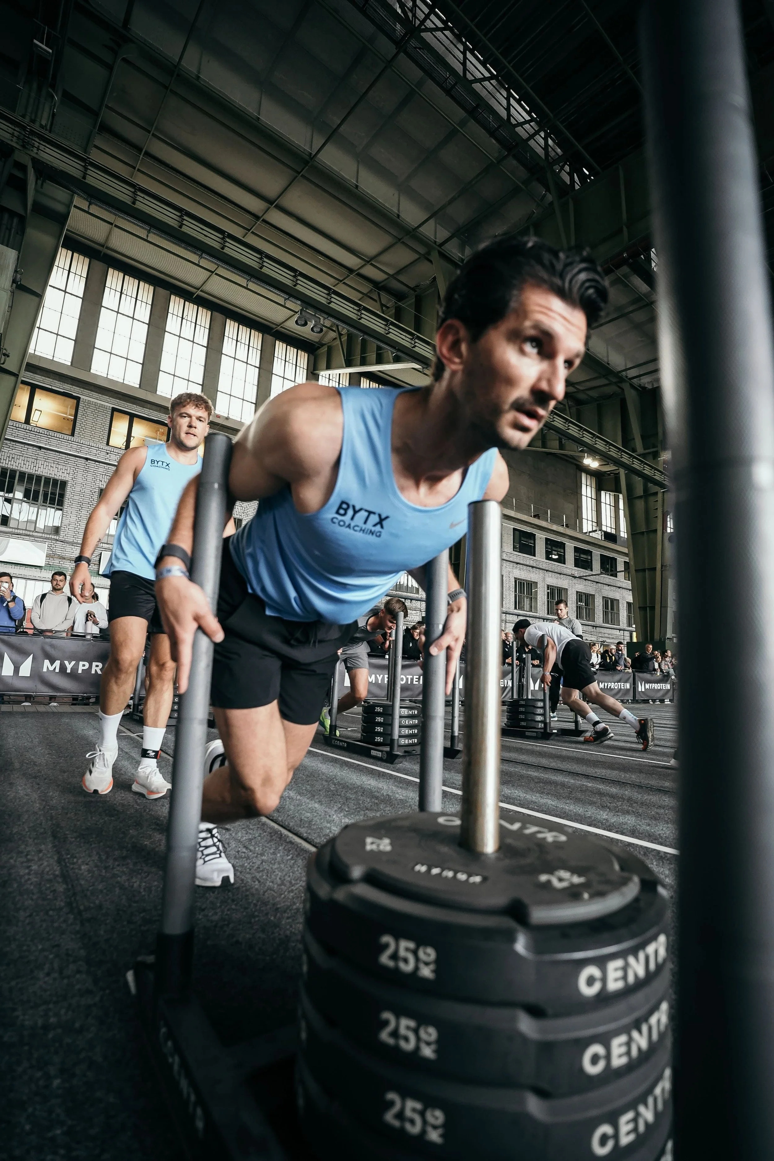 Men participating in a strength training competition inside a large industrial-style gym, pushing a weighted barbell with 25 kg weights during a workout session.