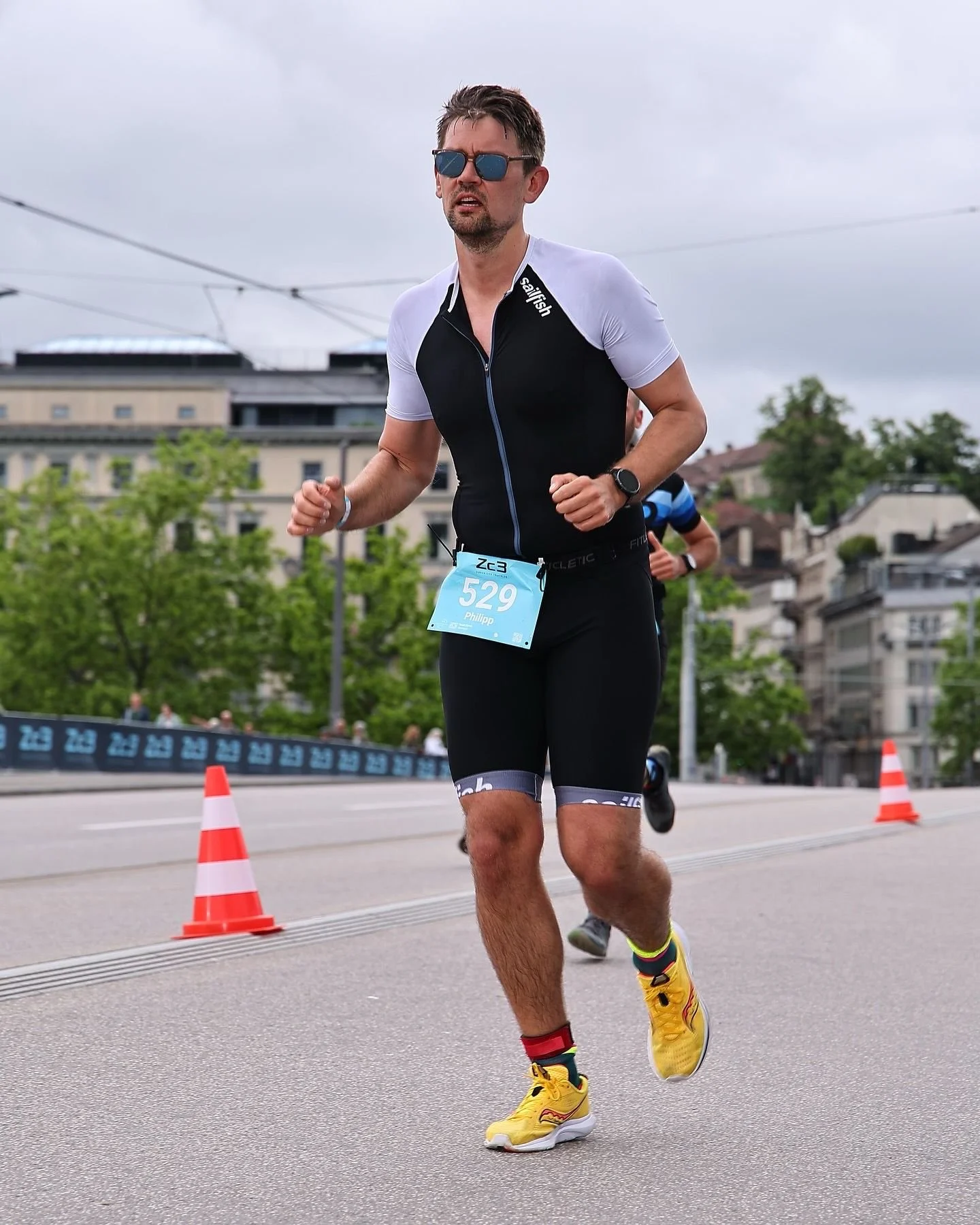 Male runner in black and white athletic outfit with yellow shoes, wearing sunglasses, participating in a zurich triathlon sprint on an urban street with orange cones and buildings in the background.