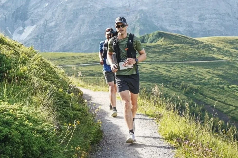 Two runners on a trail in a lush, mountainous landscape with green hills and rocky cliffs in the background.