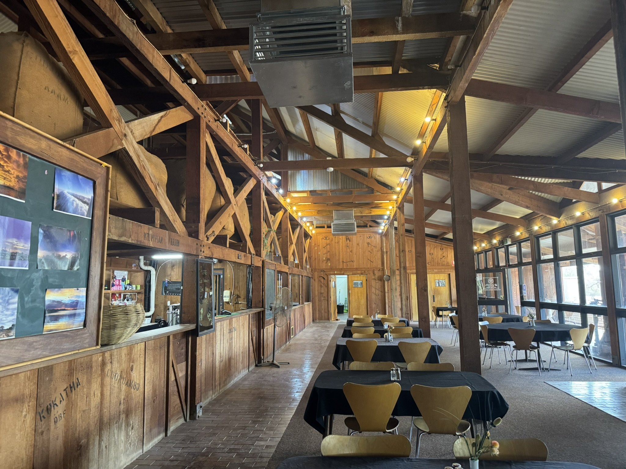 Interior of a rustic restaurant or café with wooden walls and ceiling, string lights, tables with black tablecloths and yellow chairs, large glass windows, and a serving counter with photographs on a bulletin board.