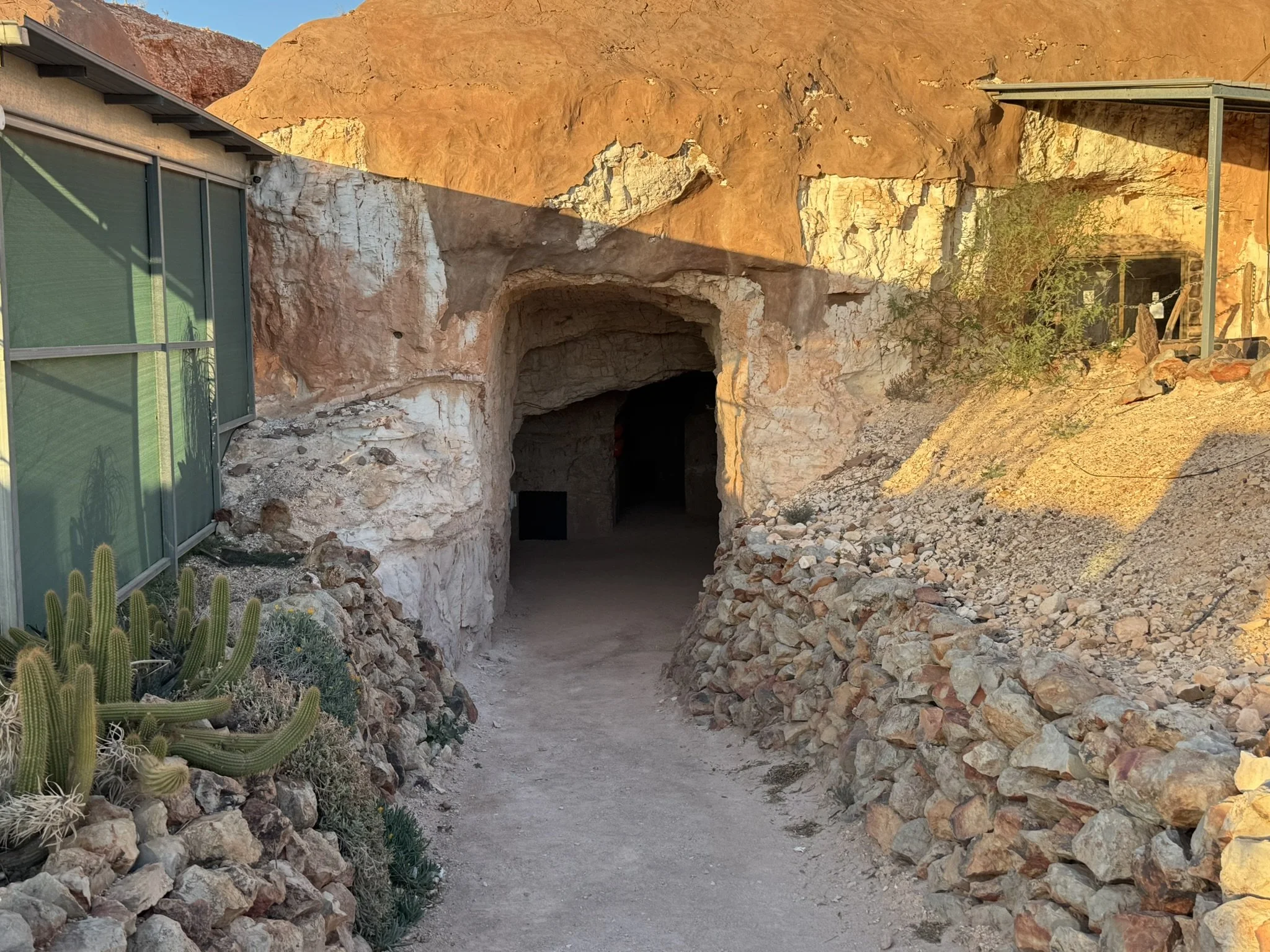 A desert pathway leading to a cave entrance in a rocky hillside, with cacti and desert vegetation on the left and a small structure with a metal roof on the right, bathed in warm sunlight.