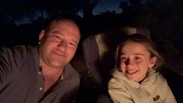 A man and a young girl smiling for a selfie outdoors at night, with trees in the background.