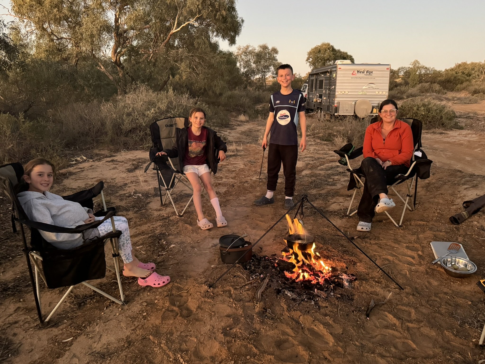 A family of five sitting around a campfire in a desert landscape during sunset, with trees and a camper in the background.
