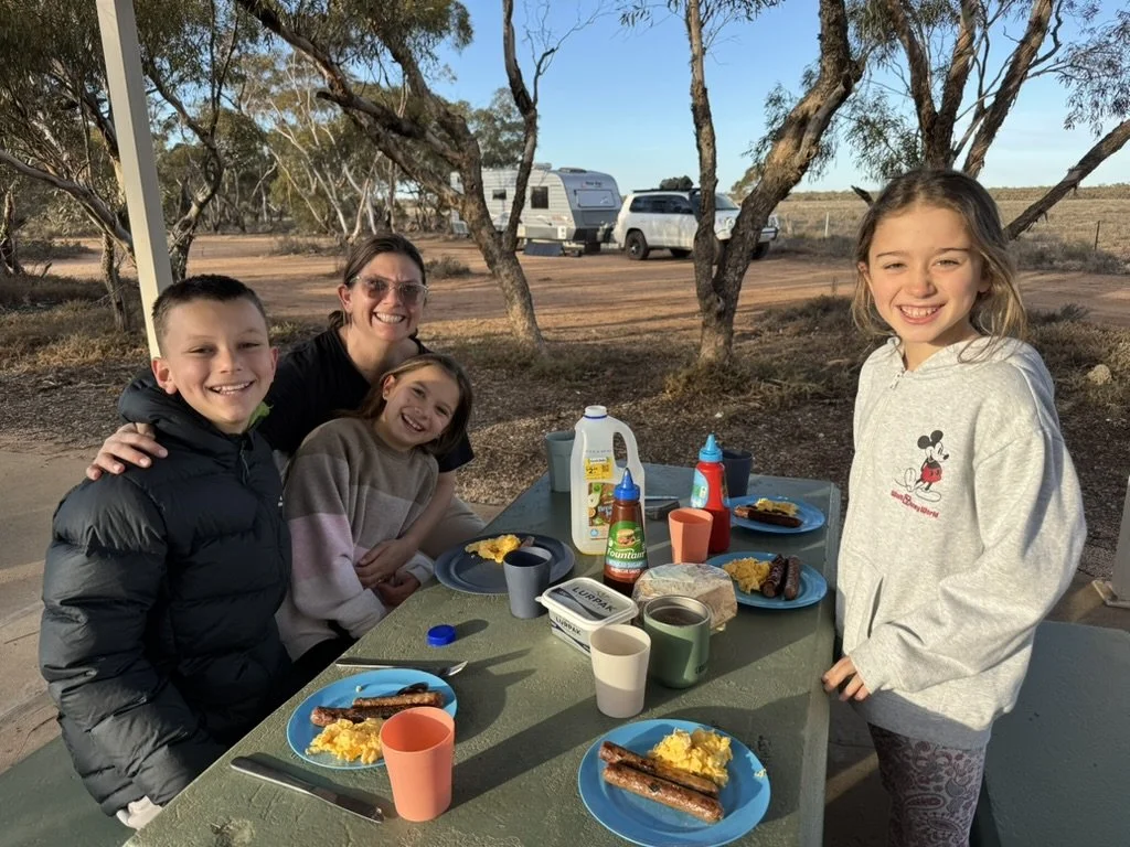 A family of five having breakfast outdoors at a picnic table with trees and parked cars in the background. The table has plates of scrambled eggs, sausages, and cups, with condiments and a bottle of syrup.