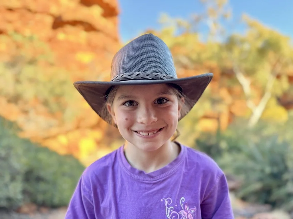 A young girl with a big smile wearing a cowboy hat and a purple shirt outdoors in front of colorful autumn trees.