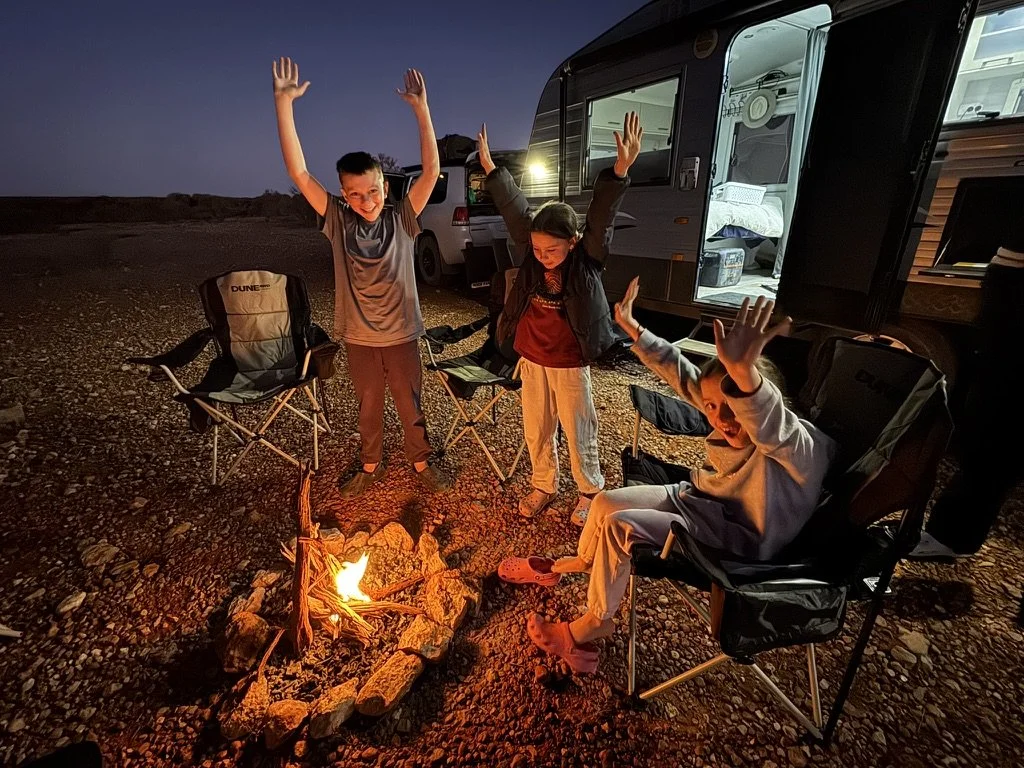 Three children around a campfire at night next to a camper trailer, smiling and raising their hands, with camping chairs nearby.