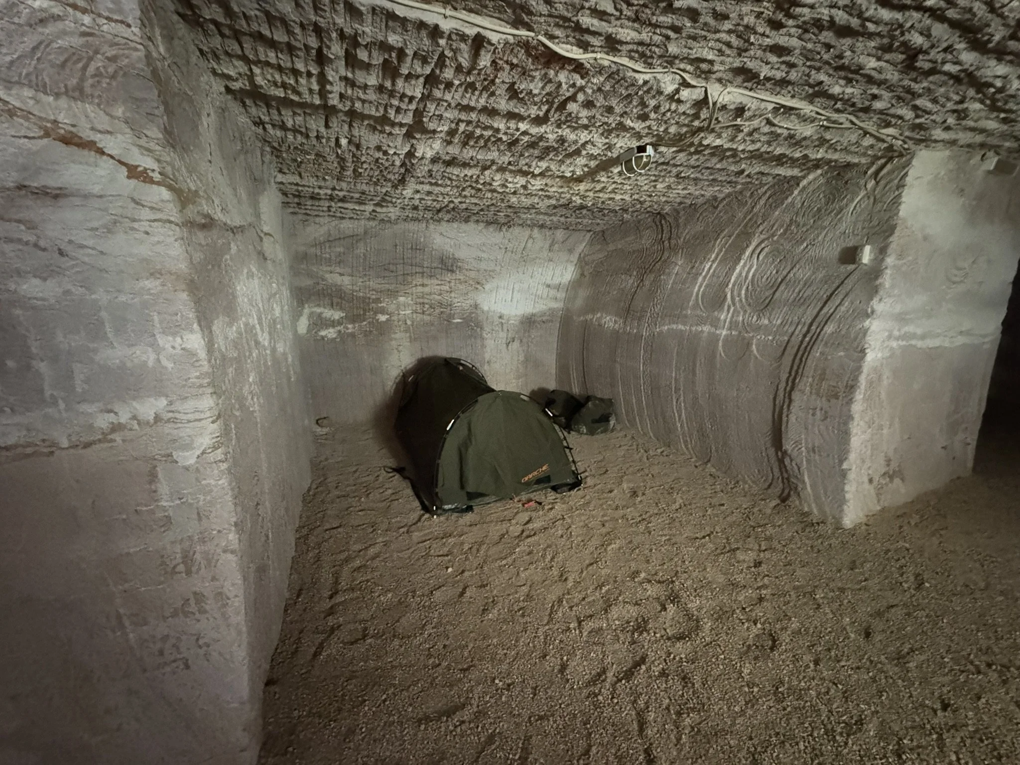 Underground tunnel with textured stone walls and ceiling. A green backpack and a small black bag are on the dirt floor.