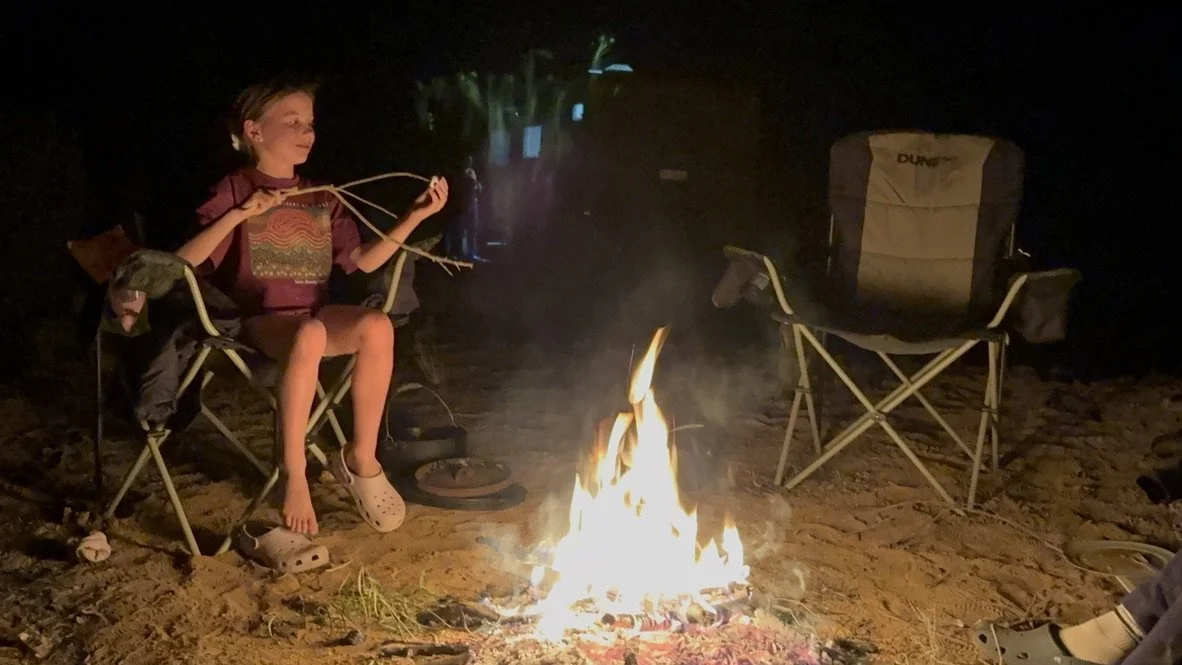 Child sitting on a camping chair by a campfire at night, holding a stick or rope, with an empty chair nearby on sandy ground.