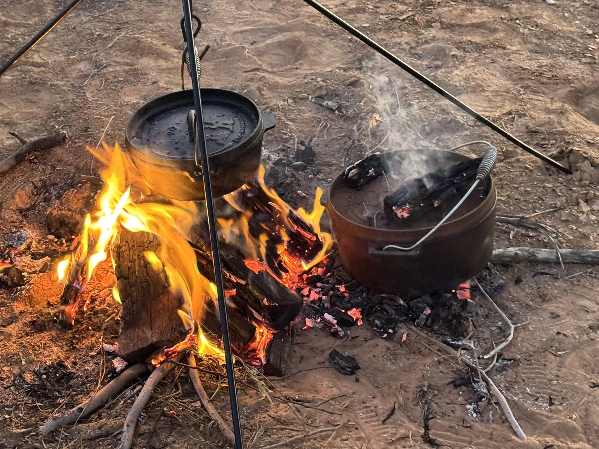 Two black pots on a campfire over a dirt ground, with flames and glowing embers visible, and a metal stand supporting the pots.