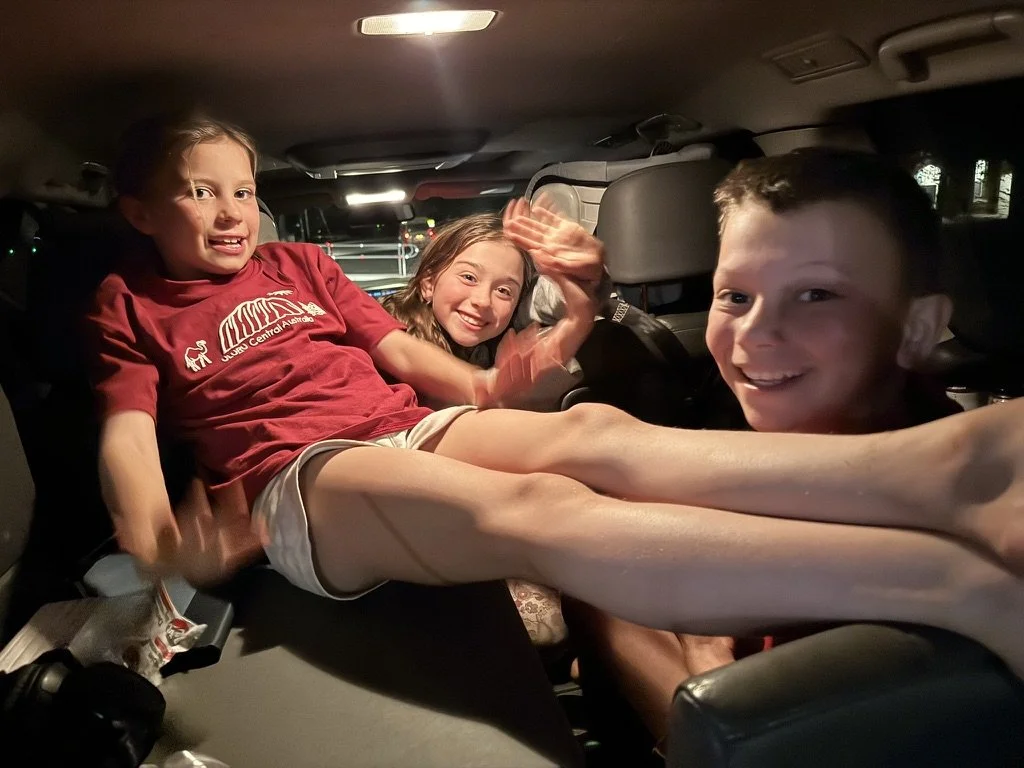 Three children sitting in the backseat of a car at night, smiling, with one girl and a boy with outstretched legs, and another girl waving.