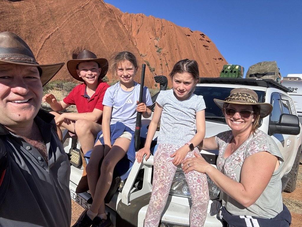 Family of five smiling and posing in front of their vehicle, with red rock formations in the background, possibly on a vacation or outdoor adventure.
