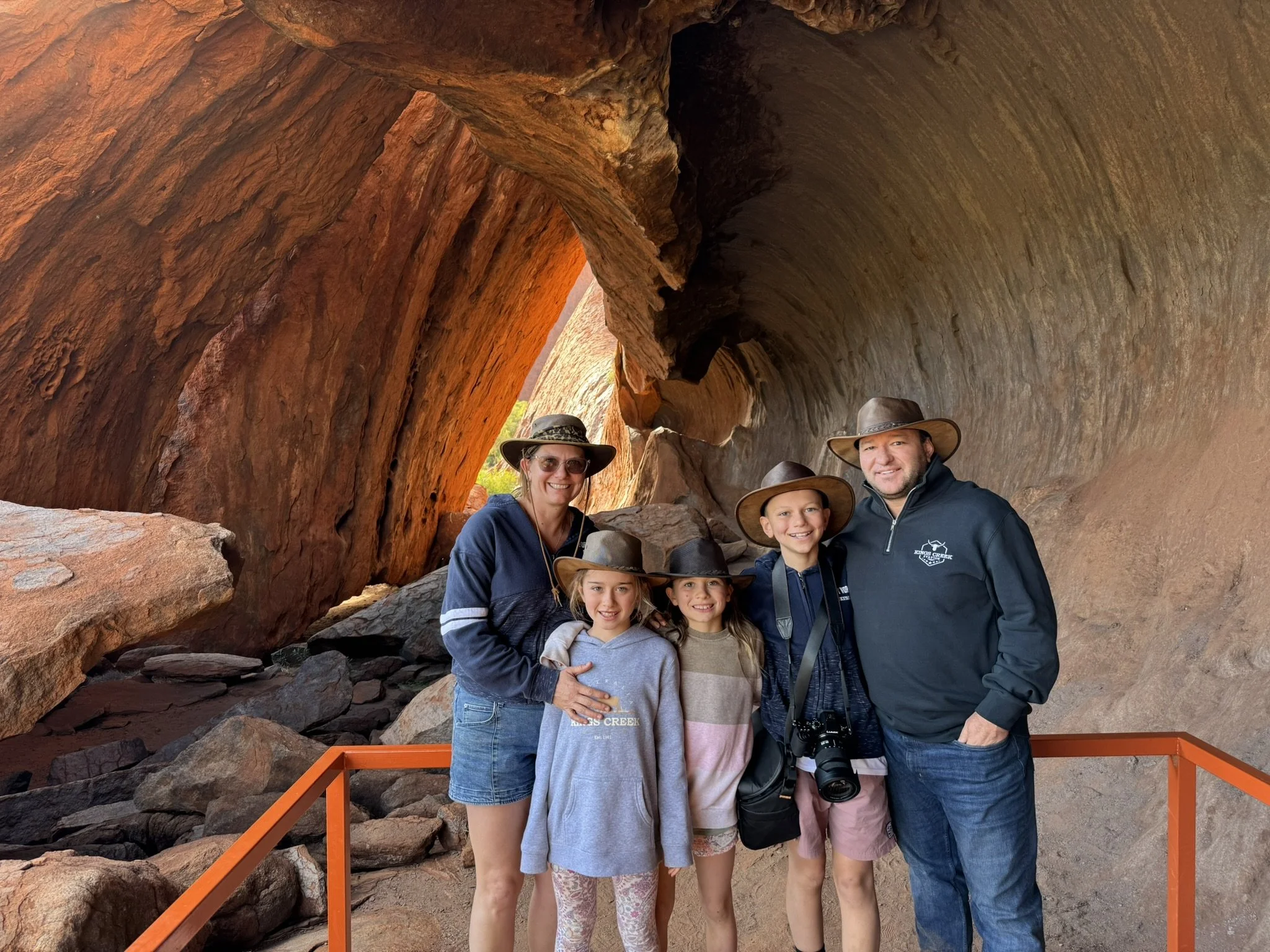 A family of five poses inside a large, curved rock formation at a canyon, all wearing hats and smiling for the photo.
